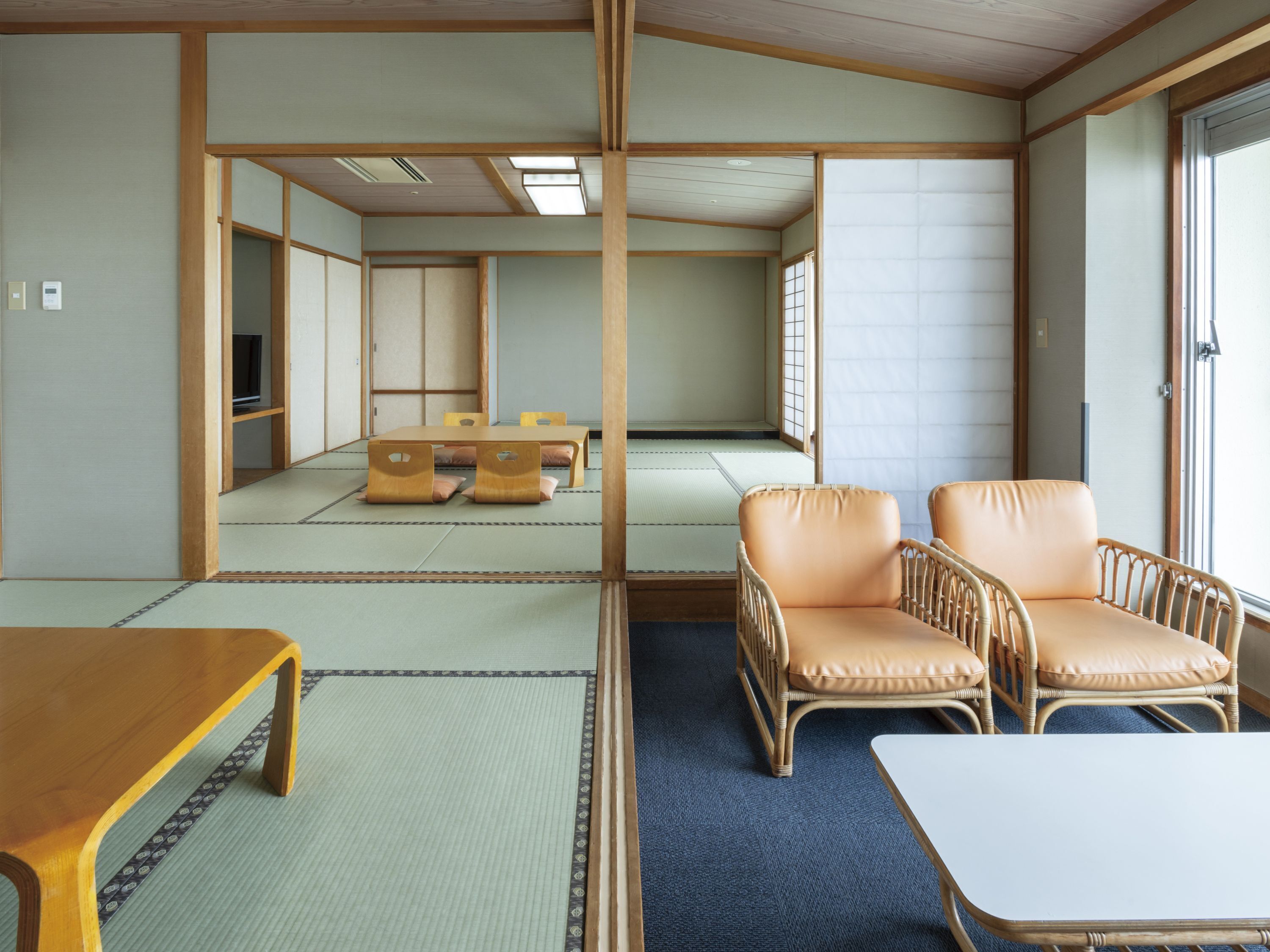 A traditional Japanese room with tatami flooring, a low table, two cushioned chairs, and a television in the background.