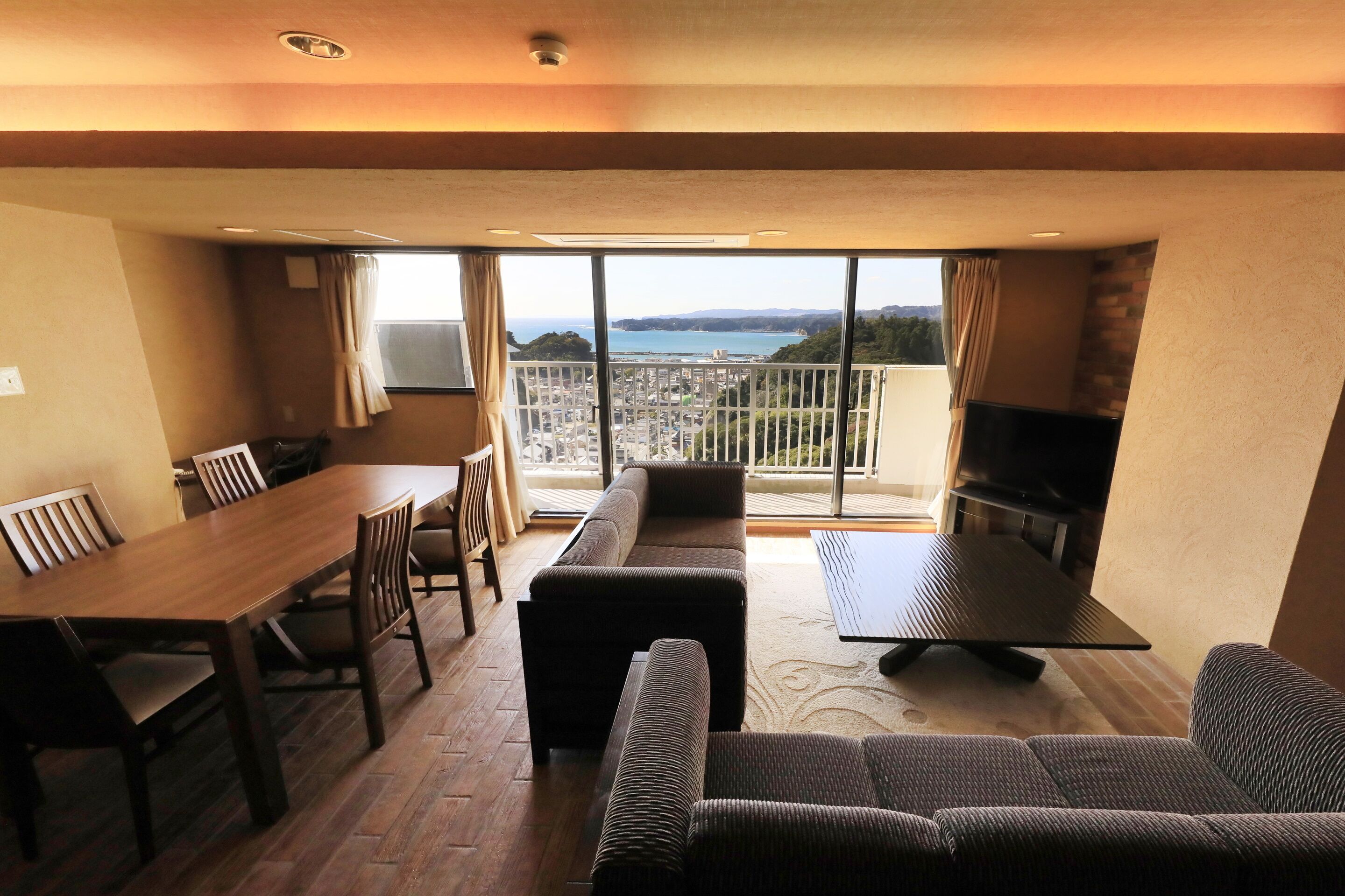 A room with a dining table, chairs, a sofa, a TV, and a balcony with a view.