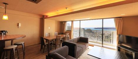 A modern hotel room with a dining area, sofa set, flat-screen TV, and a balcony with a city view.