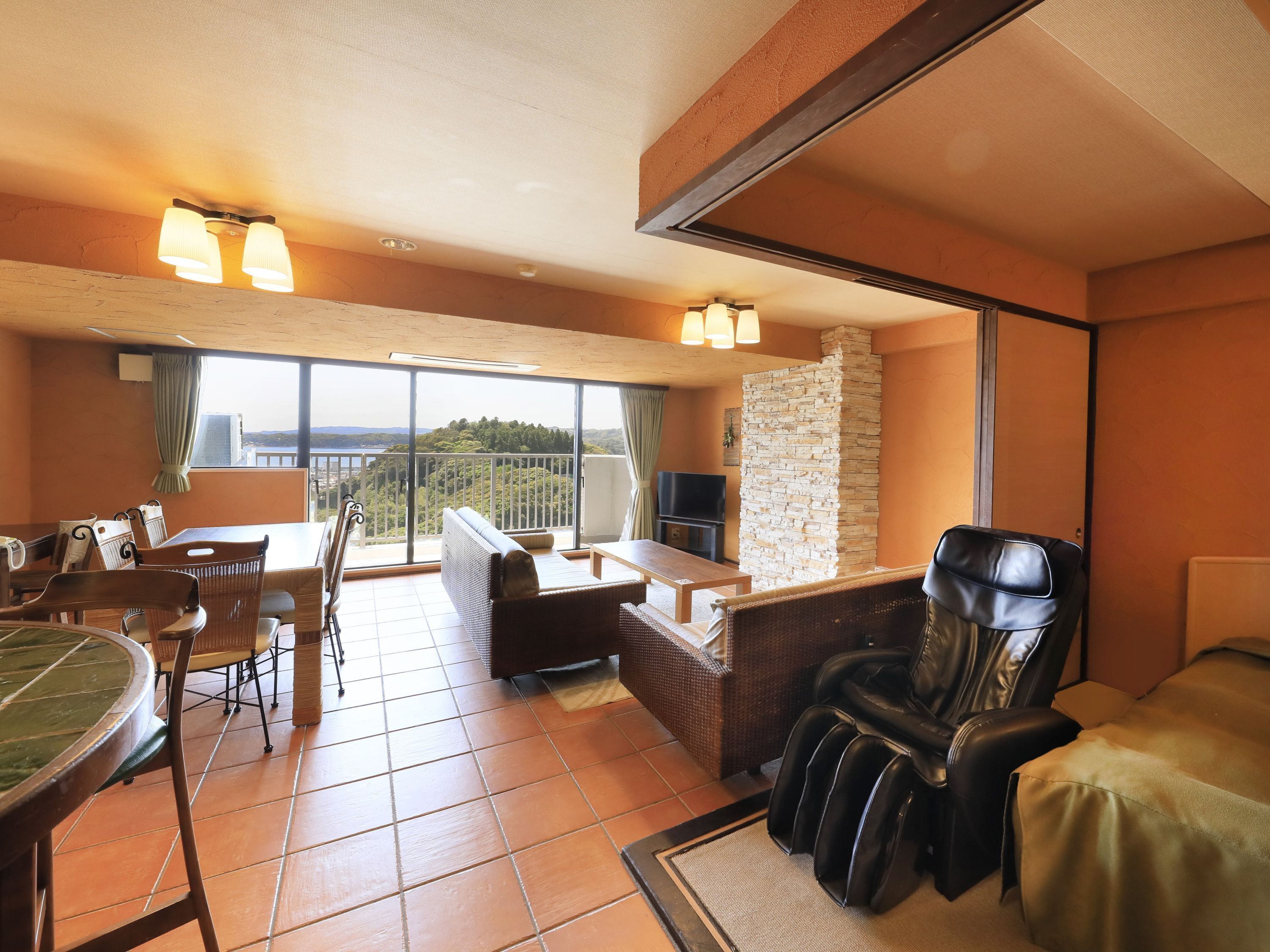Habitación de hotel con balcón, sillón de masajes, mesa de comedor y televisor.