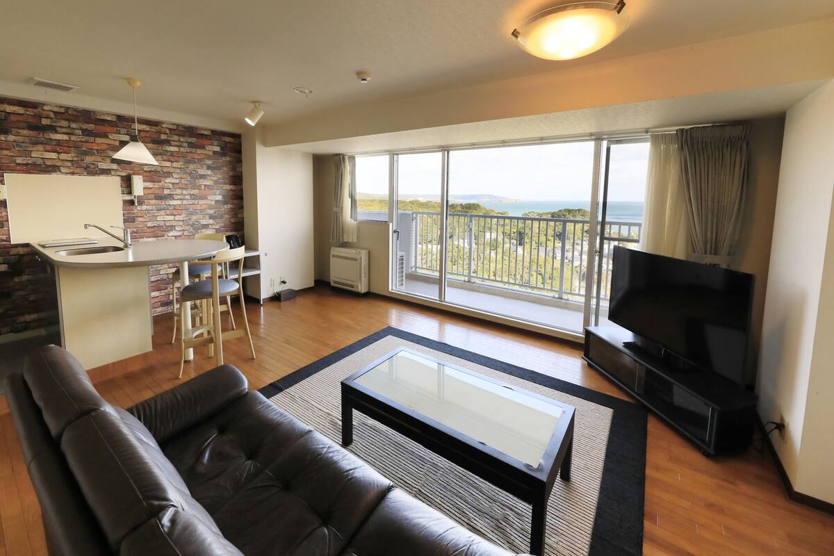 En moderne stue med en lædersofa, et glasbord, et fladskærms-tv og en stor balkon med havudsigt.
