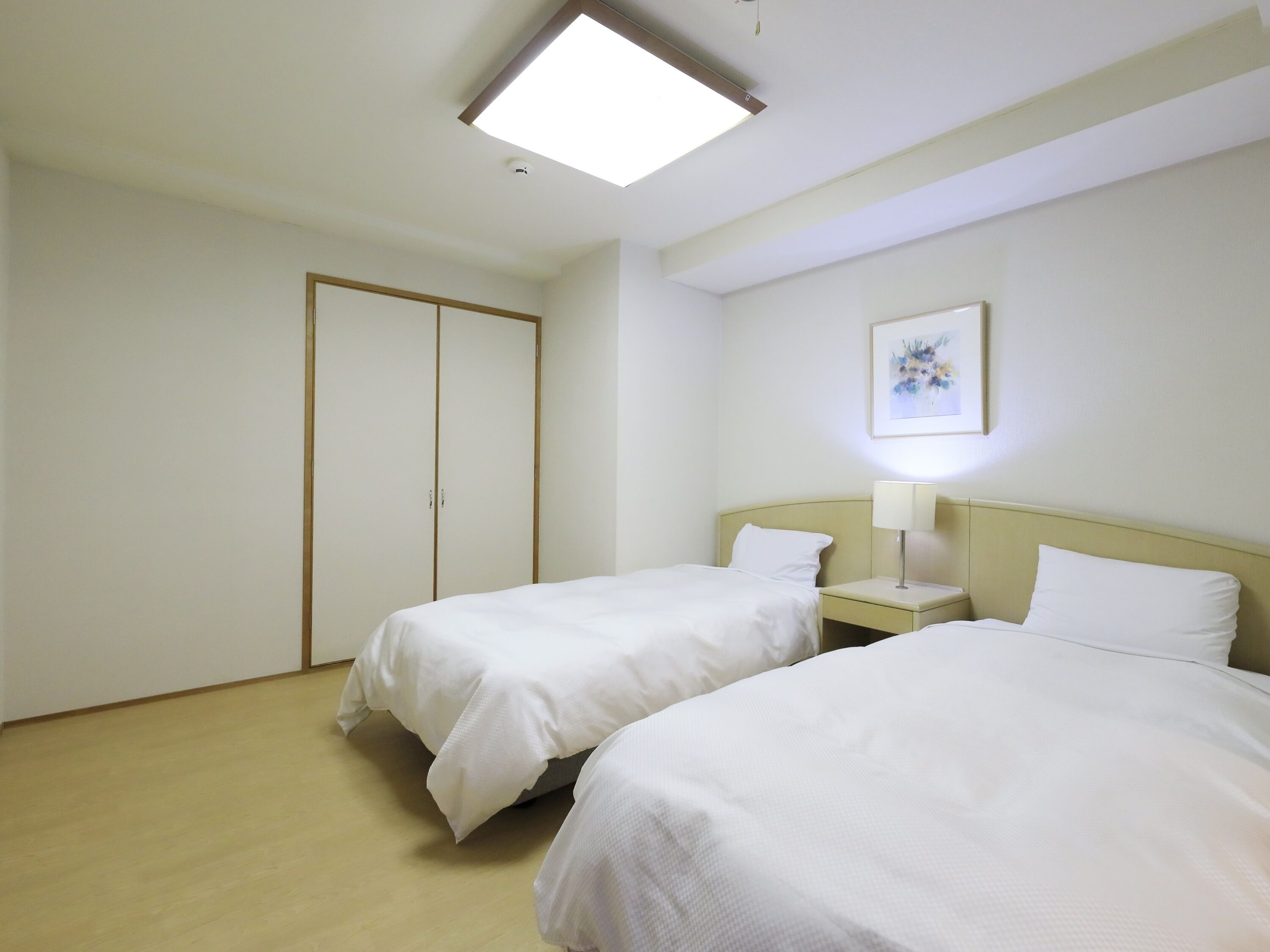 A hotel room with two beds, a bedside table with a lamp, a painting on the wall, and a ceiling light.