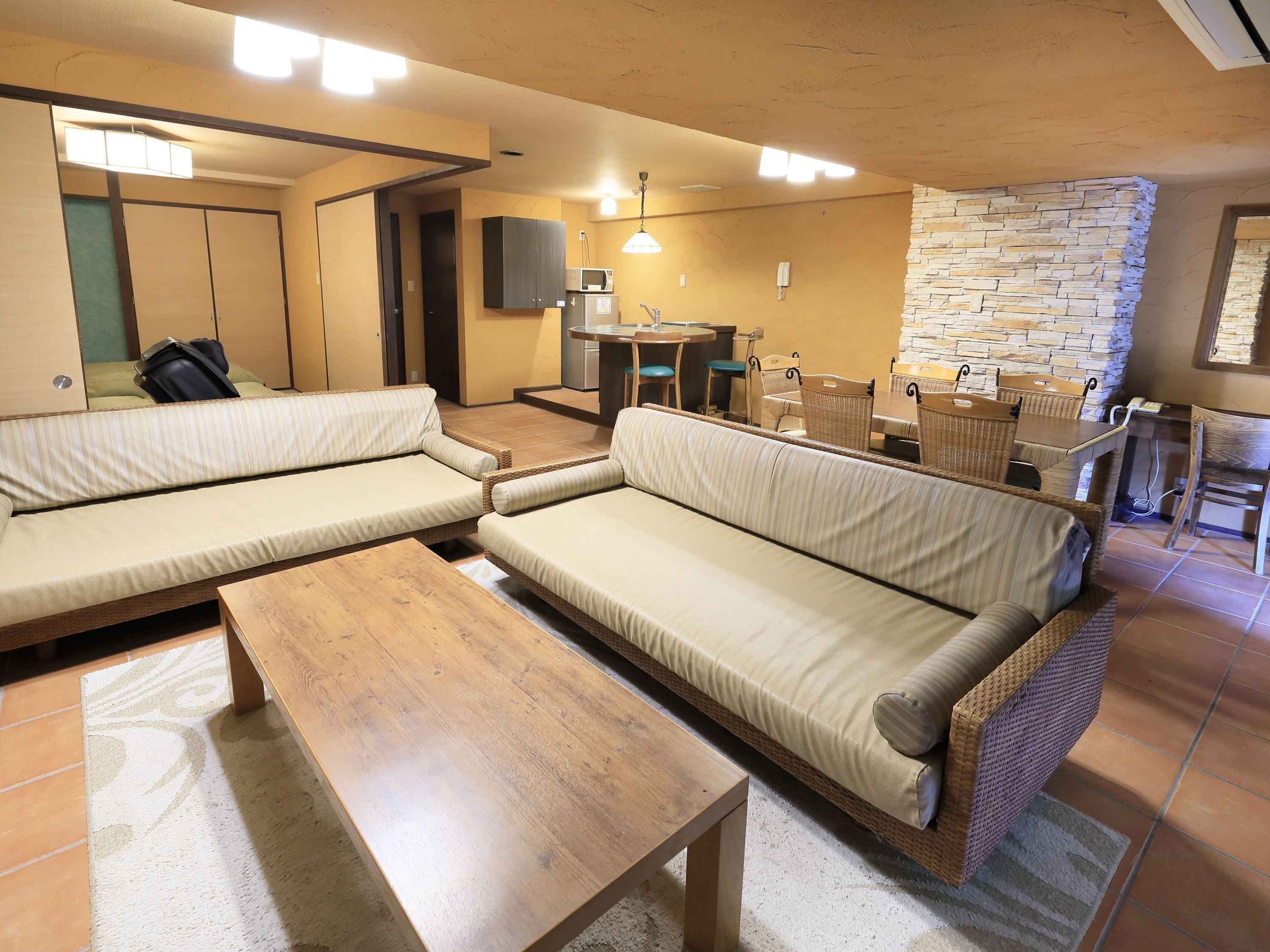 Un espace de vie compact comprenant un ensemble de canapés, une table basse en bois et un coin cuisine avec un mur en pierre.
