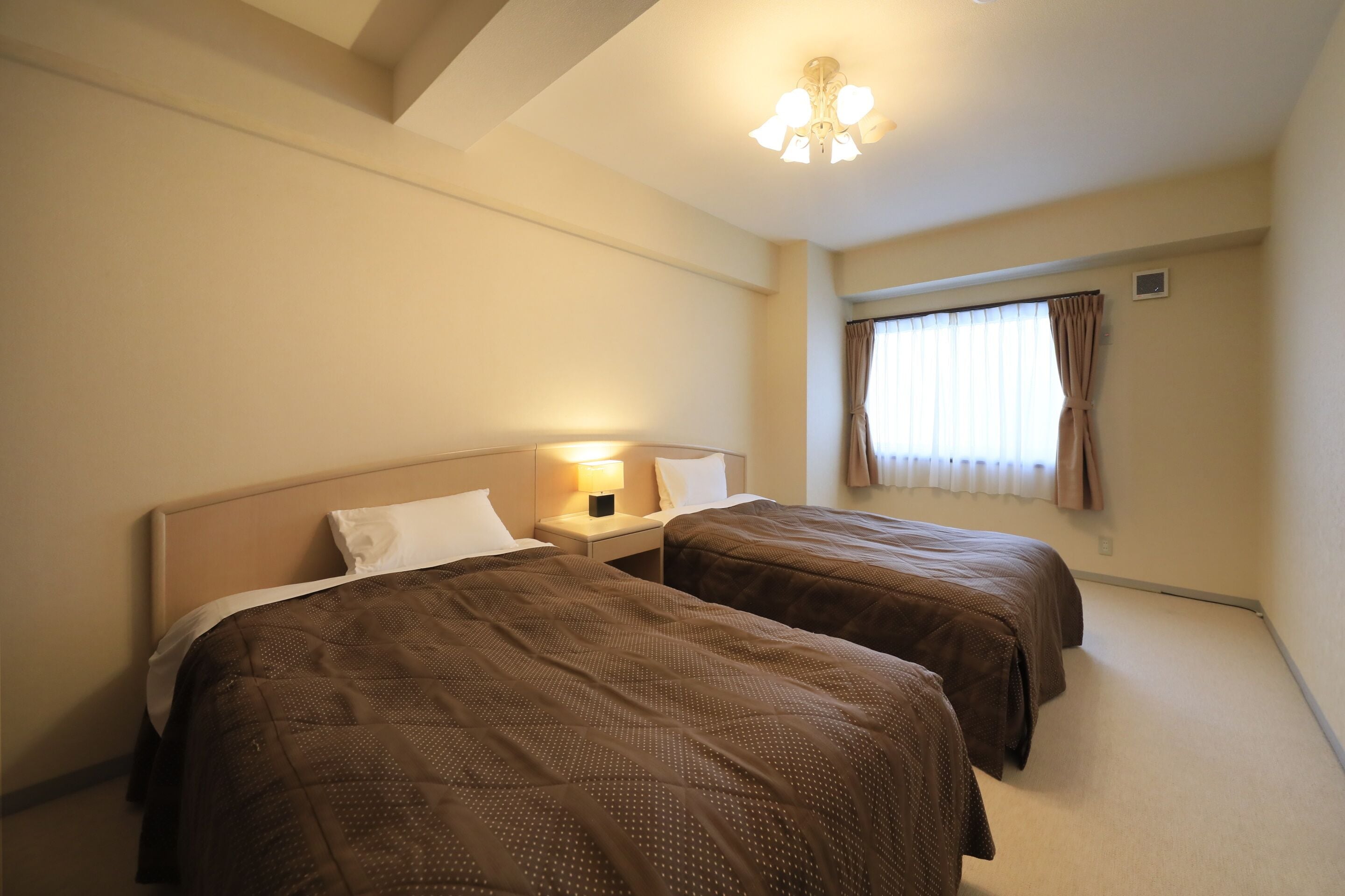 A hotel room with two beds, a nightstand with a lamp, a ceiling fan, and a window with curtains.