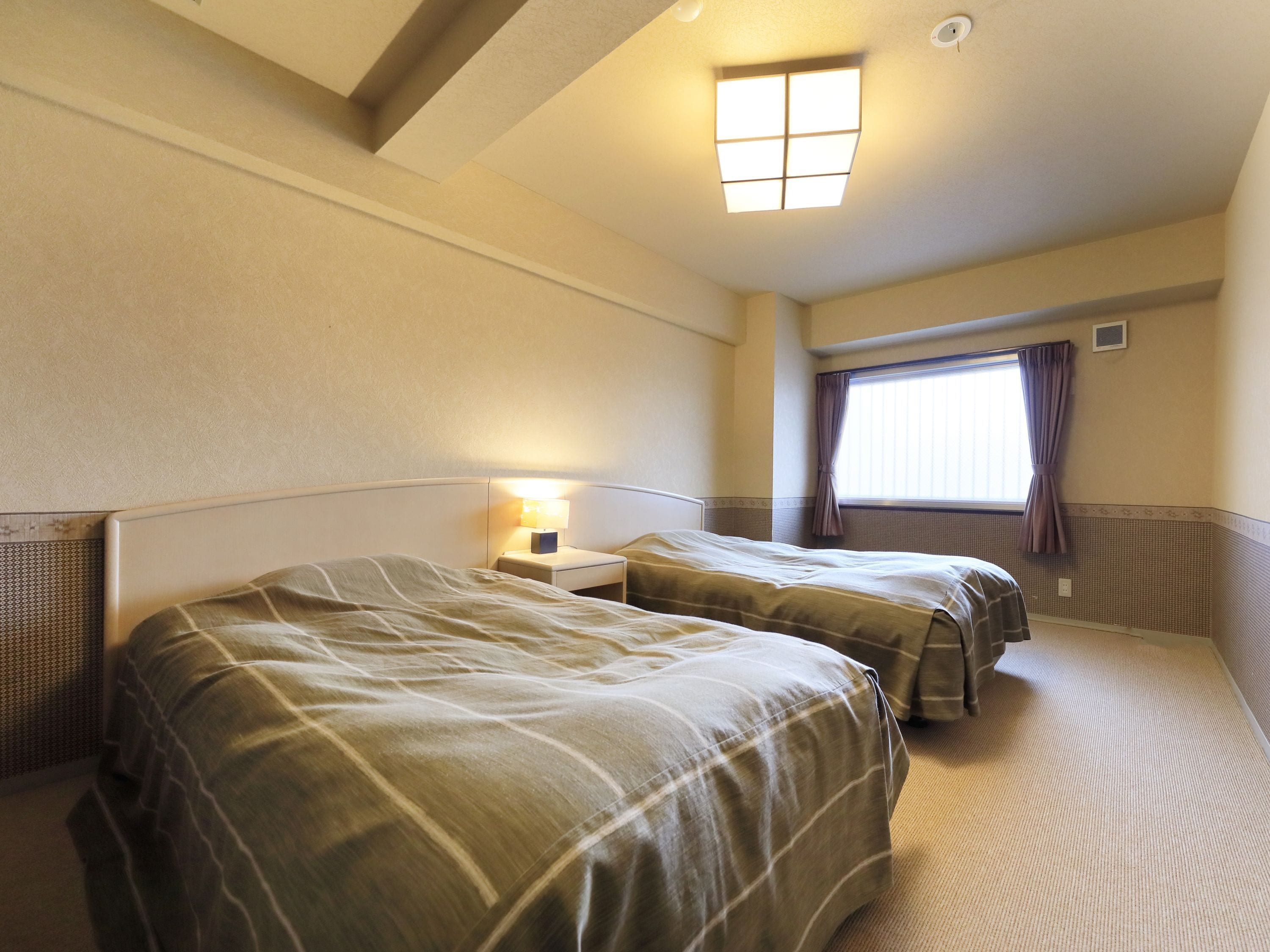 A hotel room with two beds, a nightstand with a lamp, a window with curtains, and a ceiling light fixture.