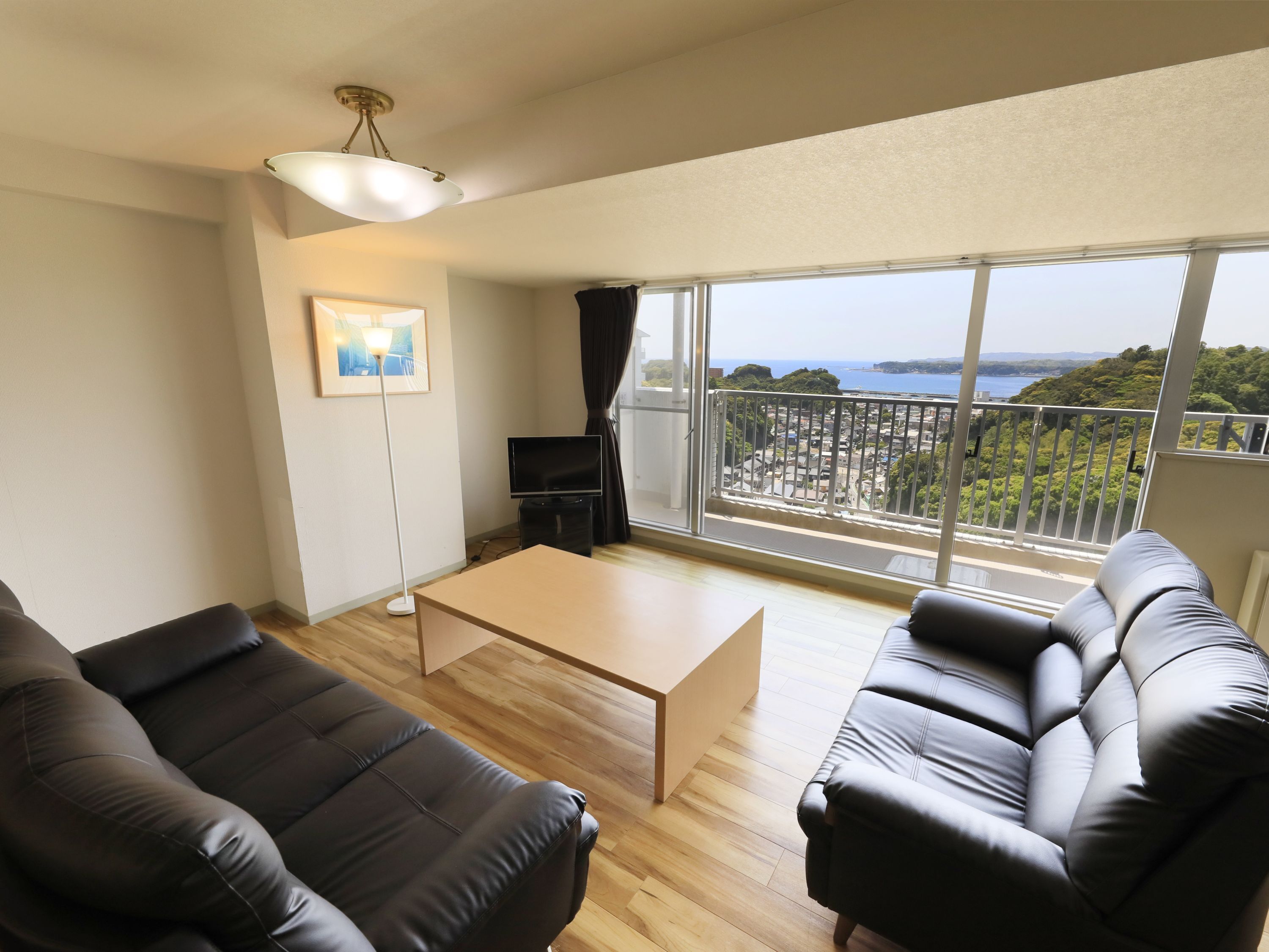 A modern living room with a sofa, a coffee table, and a TV. There is a balcony with a view of the sea and greenery.