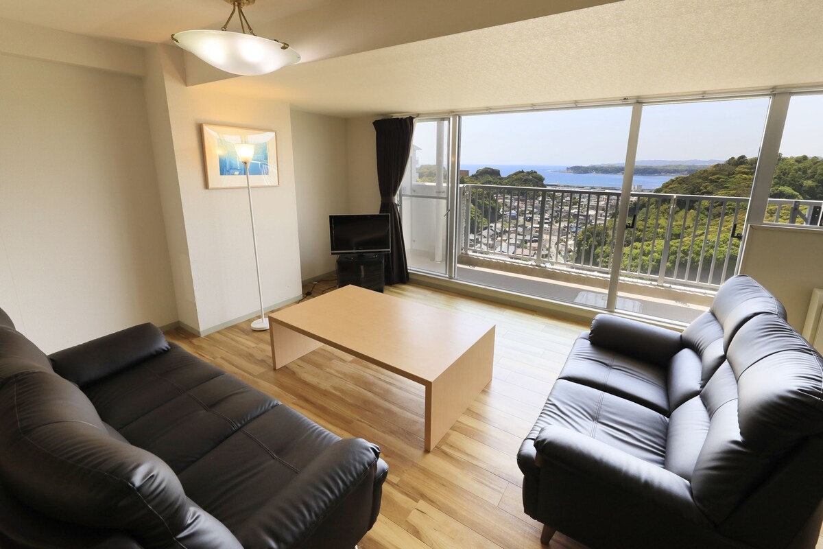 Un salon moderne avec un canapé, une table basse et un téléviseur. Il y a un balcon avec vue sur la mer et la verdure.