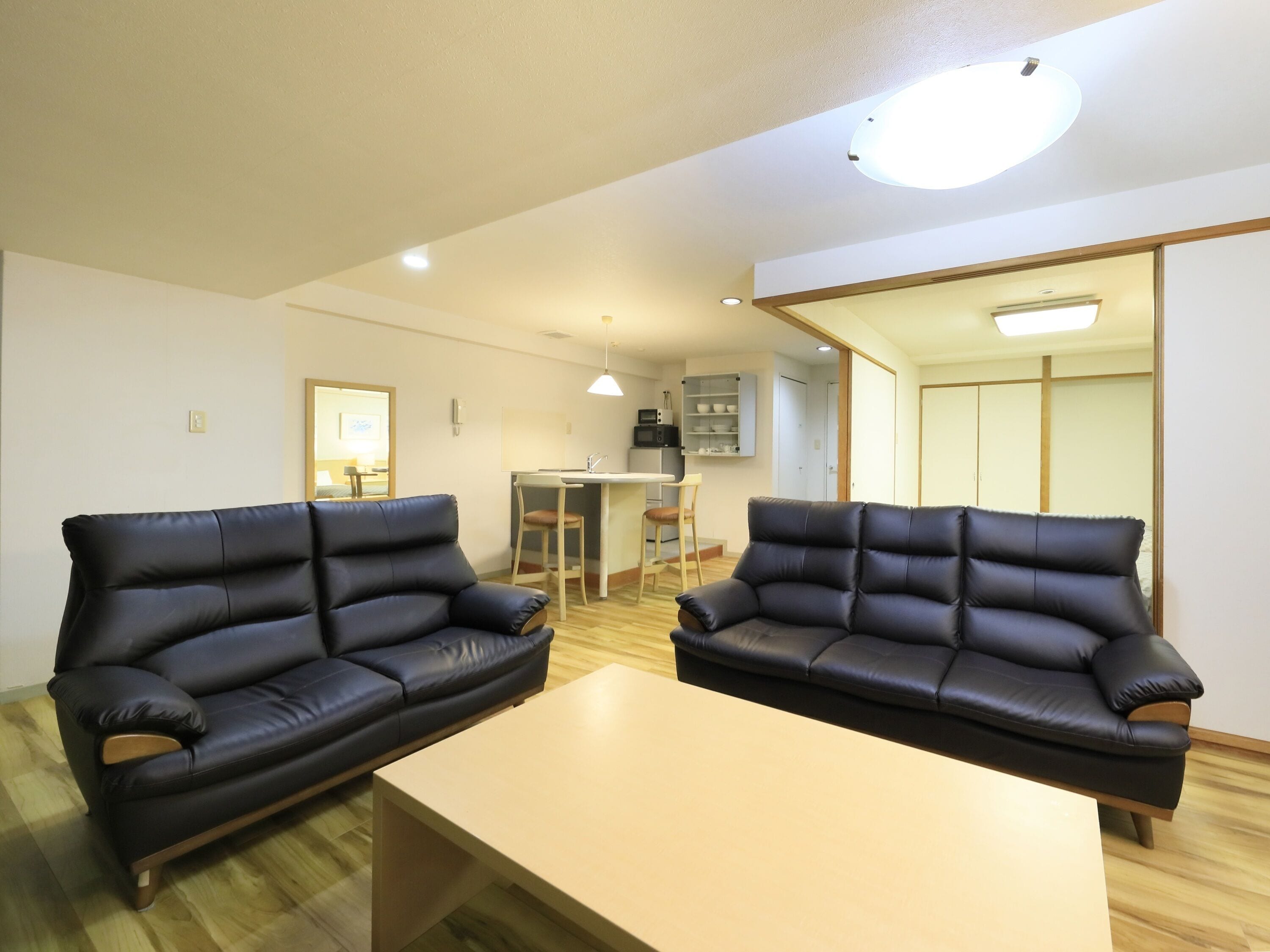 A room with two black leather sofas, a wooden coffee table, a kitchenette, and a sliding door leading to another room.