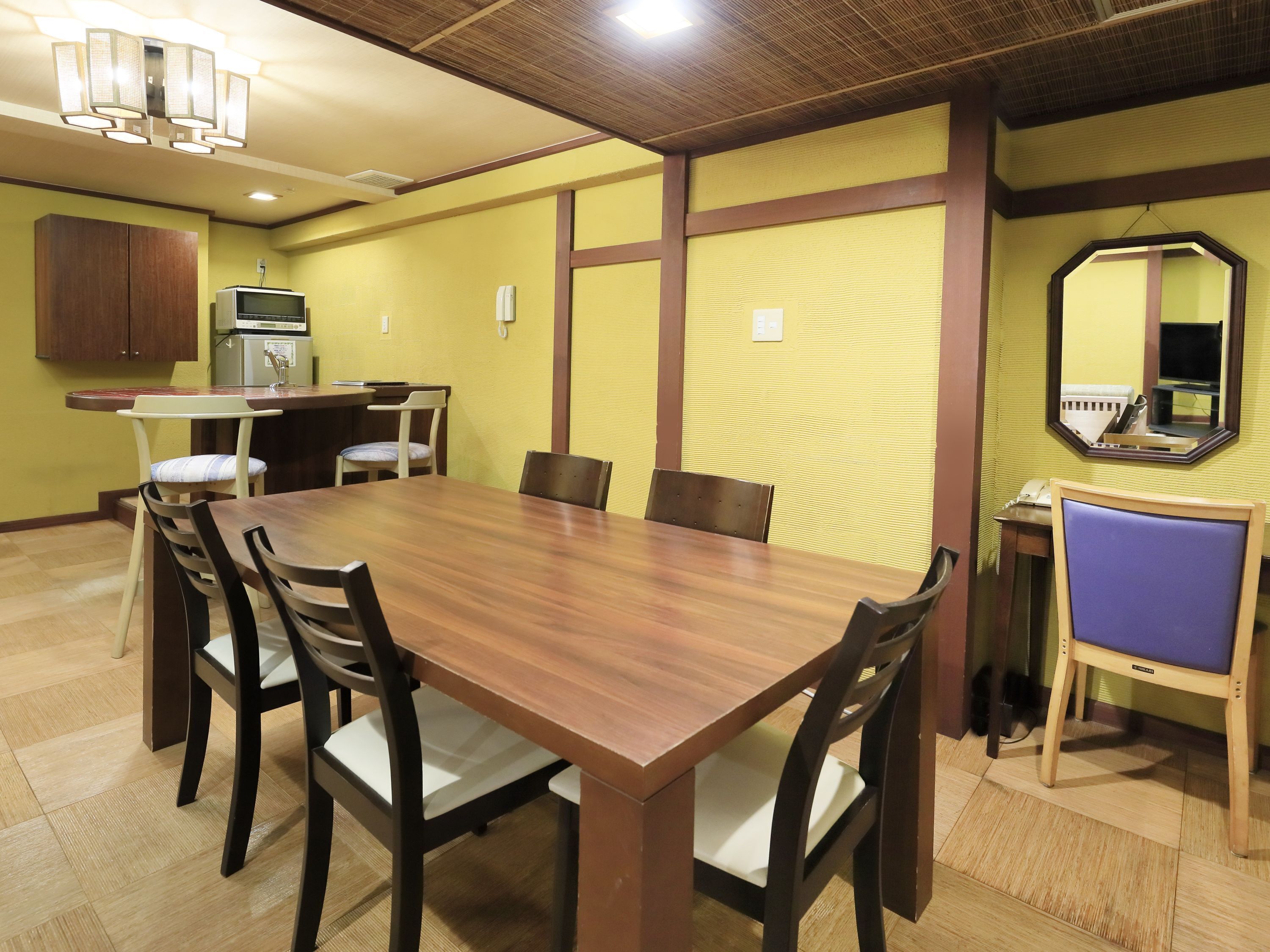 A dining area with a wooden table and chairs, a small kitchenette, and a mirror on the wall.