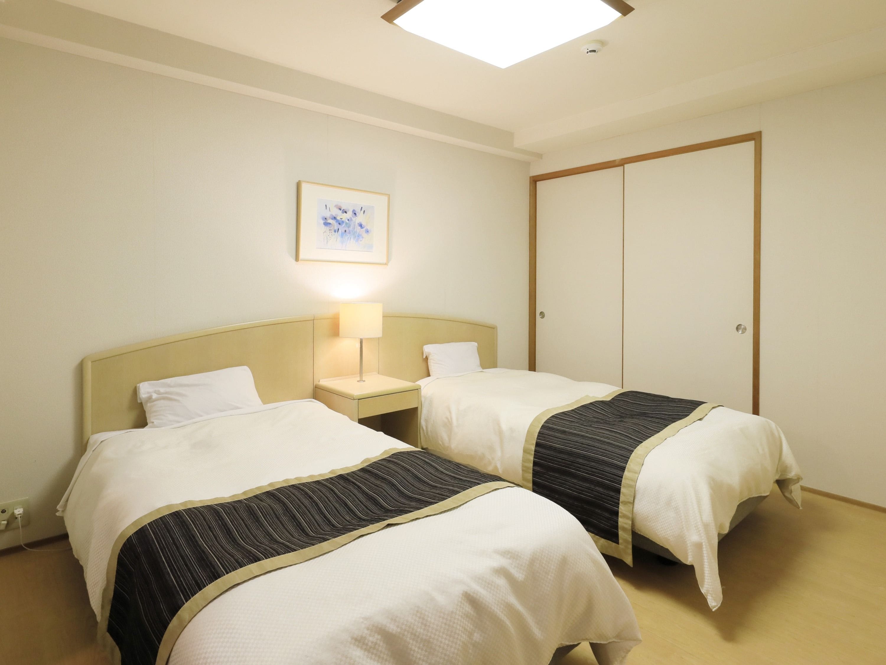 A hotel room with two beds, a bedside table with a lamp, a wardrobe, and a framed picture on the wall.