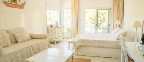 A bright, well-lit bedroom with a large bed, a sofa, a small table, and a dresser. There is a window with curtains and a view of greenery outside.