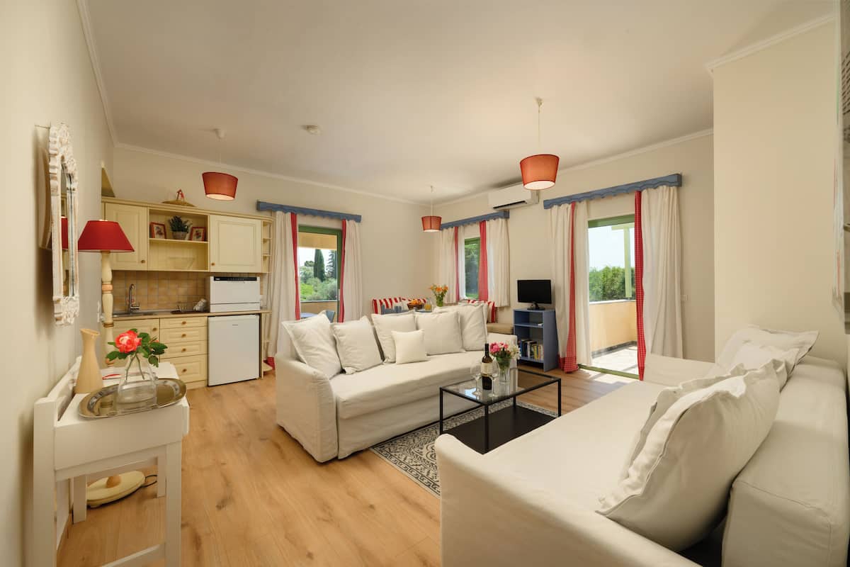 A modern living room with a sofa, a coffee table, and a TV. There is a kitchen area with cabinets and a refrigerator. The room has large windows with curtains.