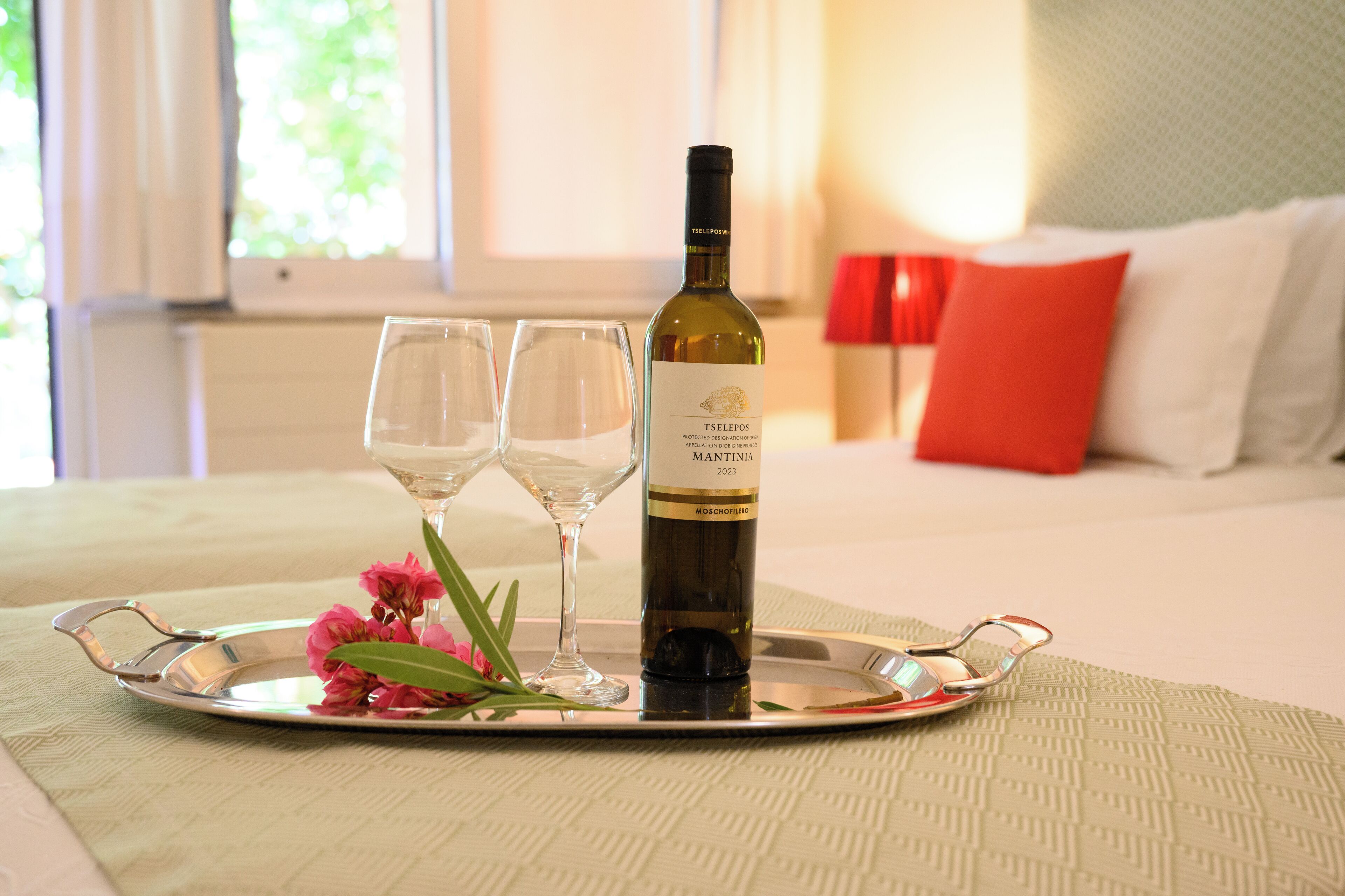 A bottle of wine on a tray with two glasses and pink flowers, set on a bed with a red pillow and a lamp in the background.