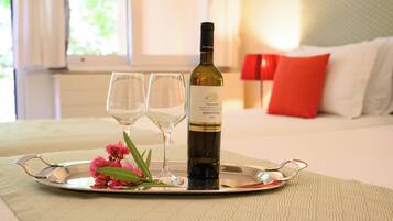 A bottle of wine on a tray with two glasses and pink flowers, set on a bed with a red pillow and a lamp in the background.