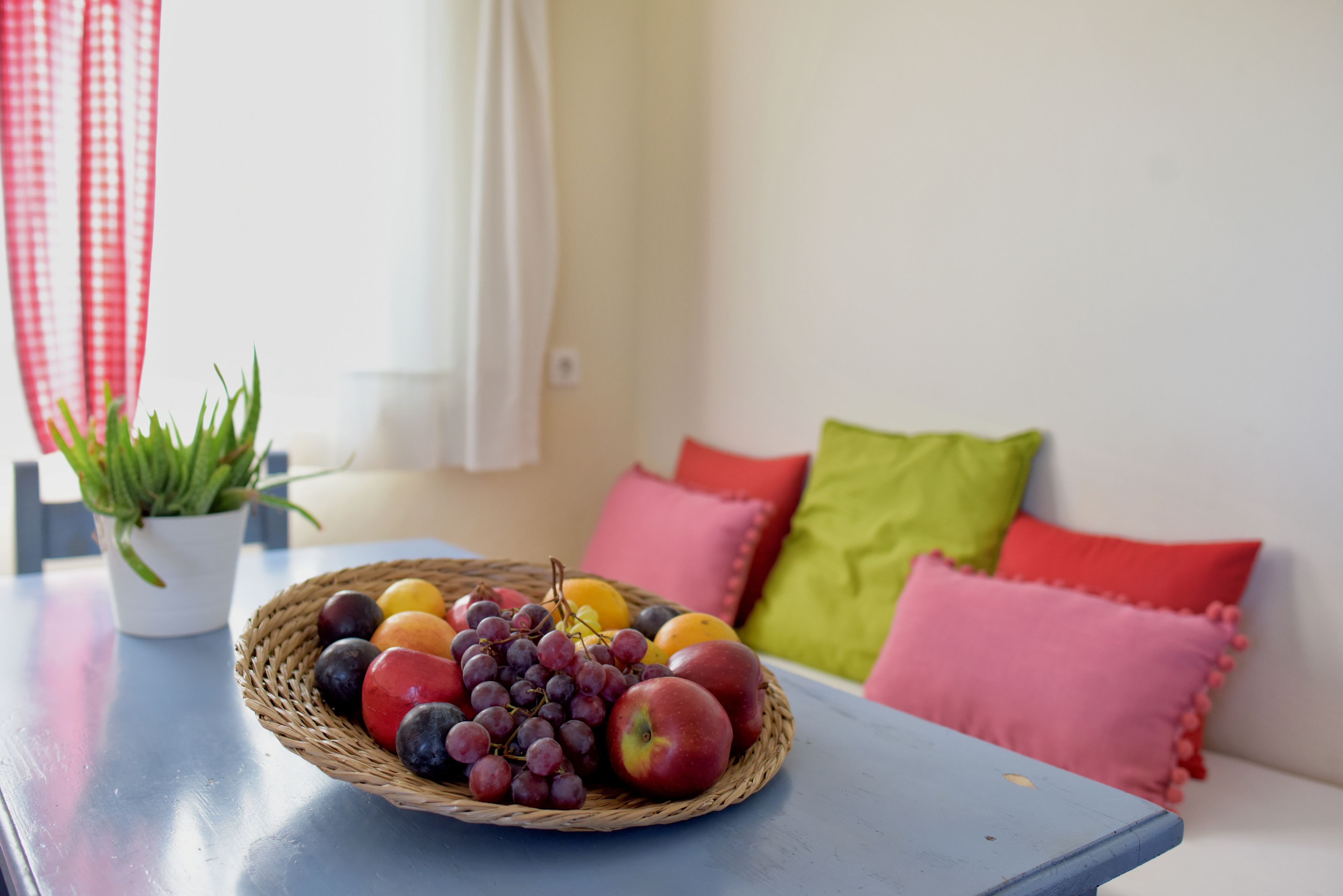 A wicker fruit basket on a blue table with a potted plant and colorful pillows in the background.