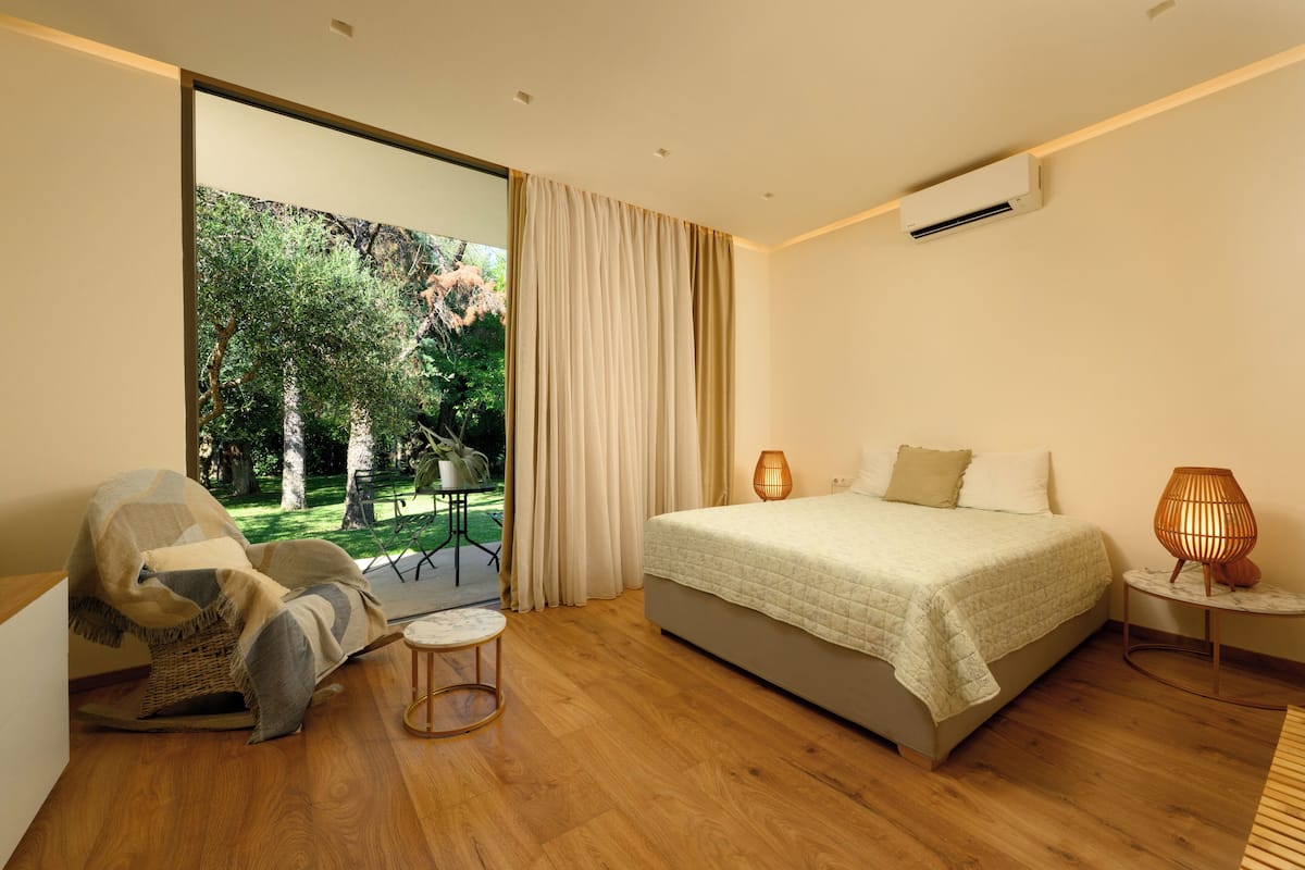 A modern bedroom with a large bed, a chair, a small table, and a view of a garden through a sliding door.