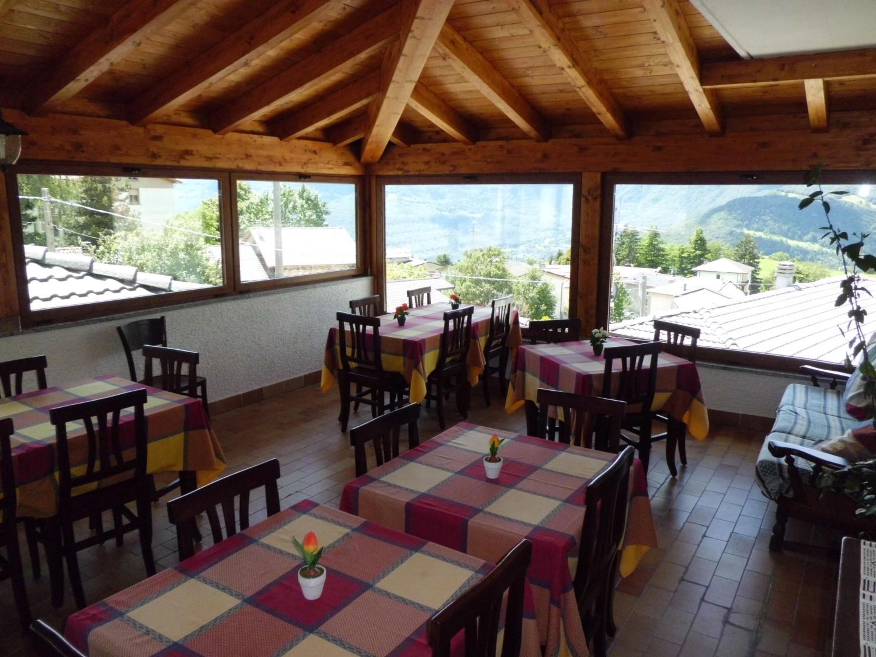 Un'area pranzo con tavoli e sedie in legno, ampie finestre vista montagna e un soffitto alto in legno.