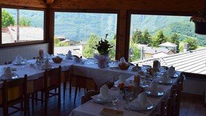 A dining area with tables set for a meal, large windows offering a view of a mountainous landscape, and wooden beams on the ceiling. - Albergo Canella (Fuipiano Valle Imagna)