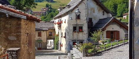 A traditional stone-paved courtyard with rustic buildings, a cobblestone street, and a mountainous backdrop.