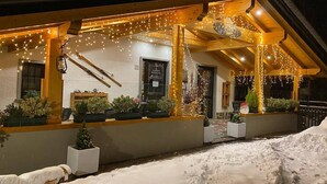 A snow-covered building with a wooden porch decorated with string lights and potted plants. - Albergo Canella (Fuipiano Valle Imagna)