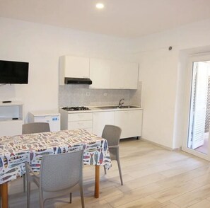 A compact kitchen with white cabinets, a gas stove, and a sink. There's a dining table with chairs and a TV mounted on the wall. - Villa Mare (Portoferraio)