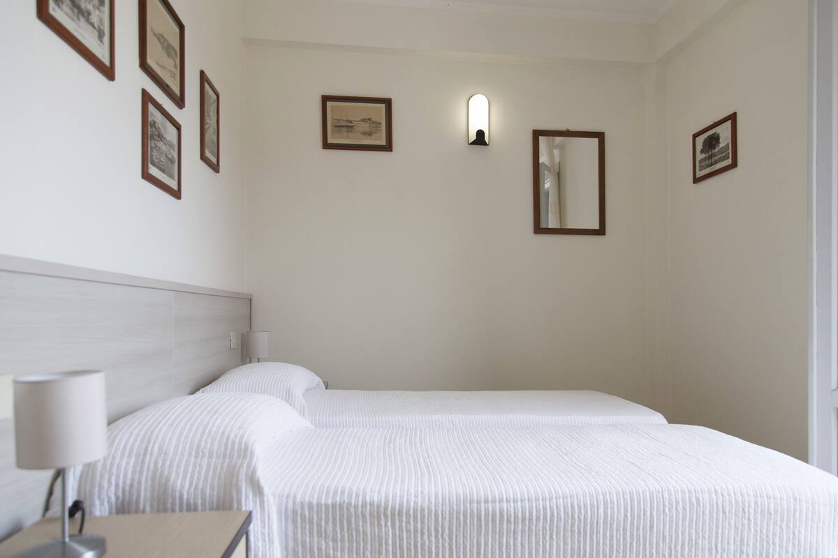 Habitación de hotel con dos camas, una mesita de noche con lámpara y cuadros enmarcados en las paredes.