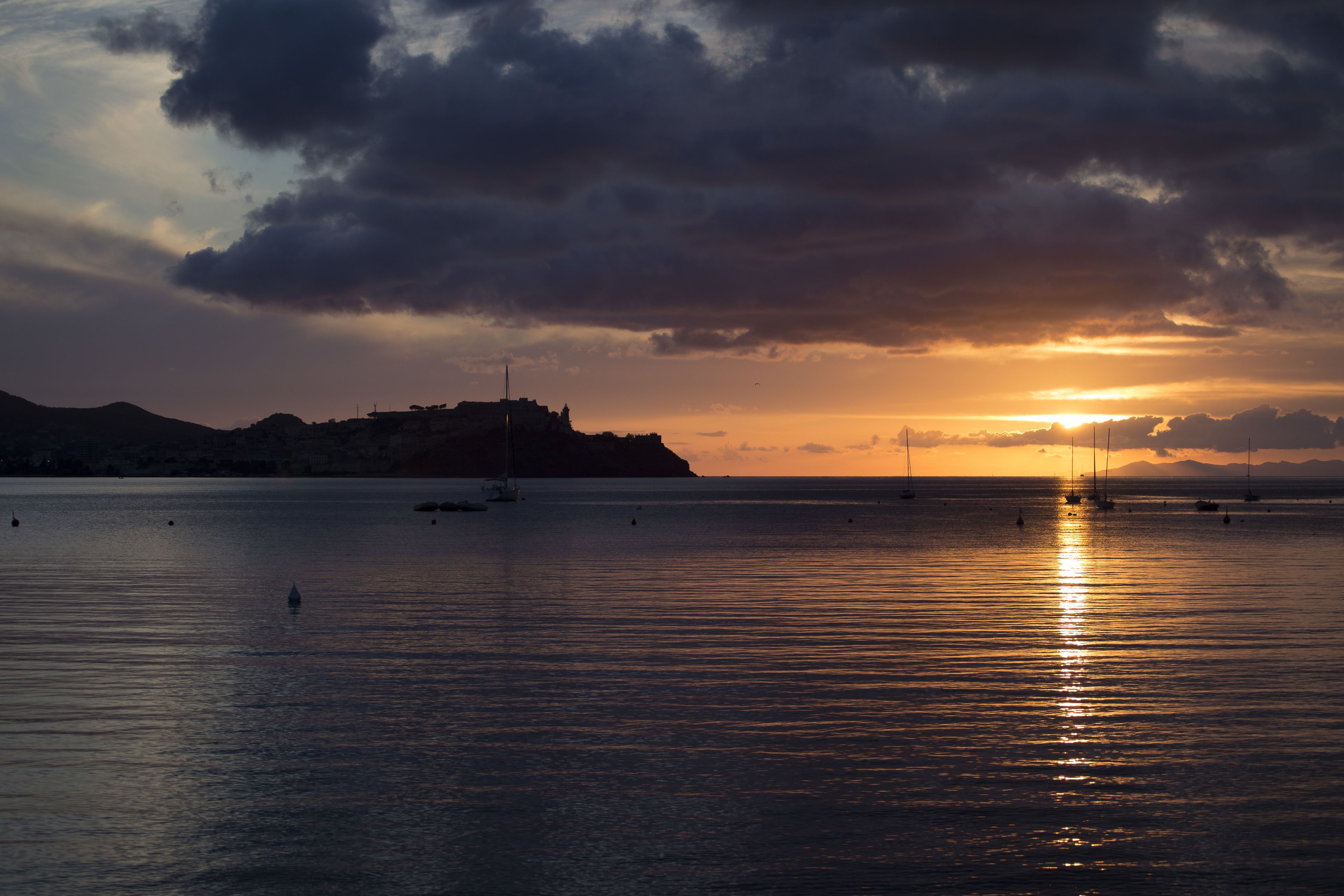 Une mer calme au coucher du soleil, avec des bateaux et une colline au loin.