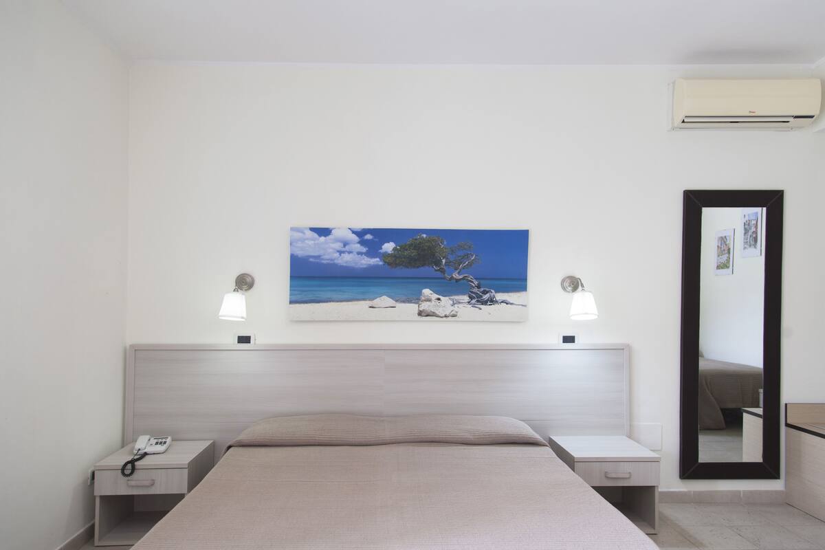 Habitación de hotel con cama, mesitas de noche, un cuadro de una playa, aire acondicionado y un espejo.