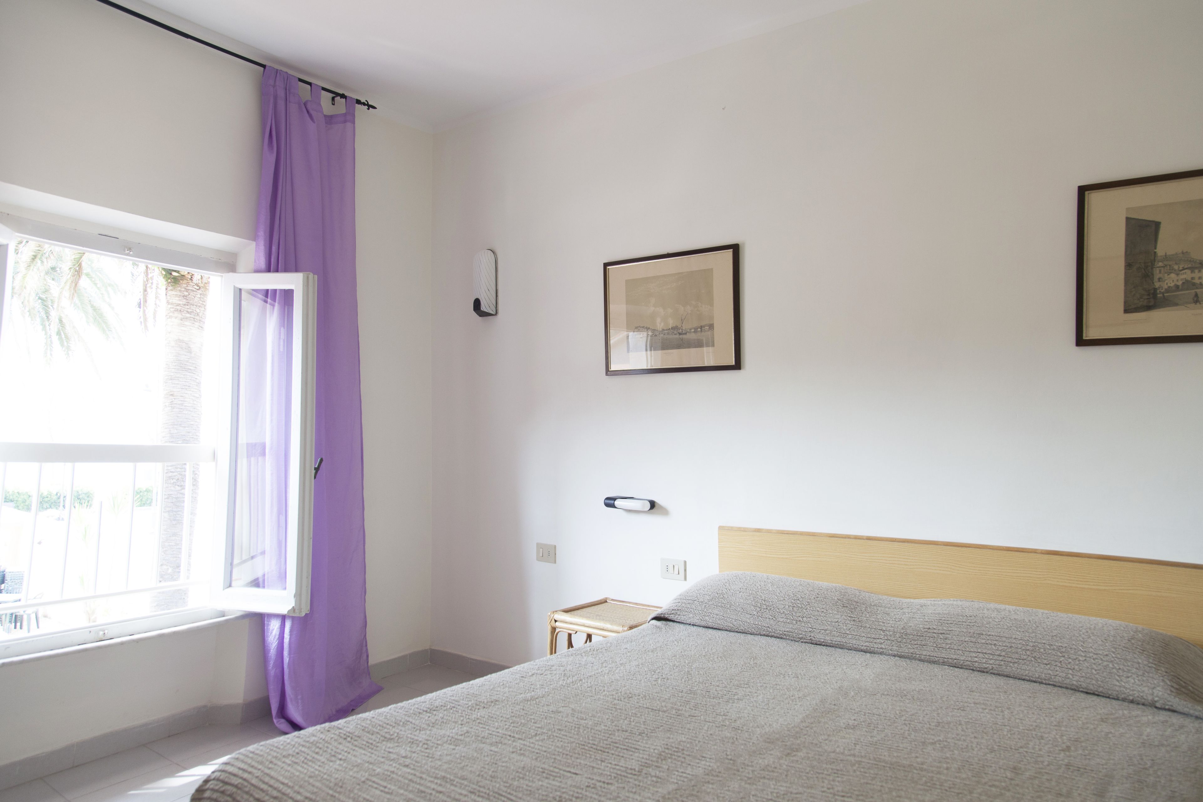A bedroom with a bed, a window with purple curtains, a bedside table, and two framed pictures on the wall.