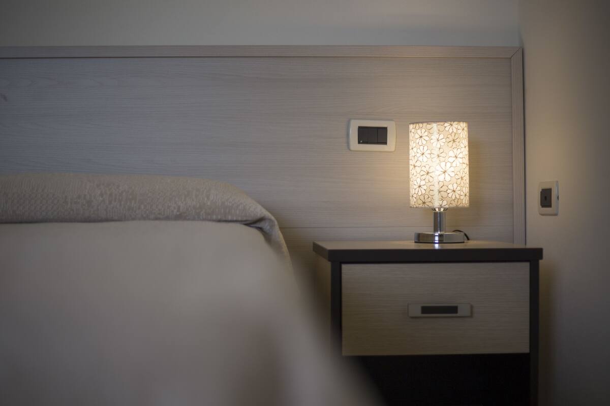 Una cama bien hecha con cabecera, una mesita de noche con lámpara y un pequeño panel de control en la pared.