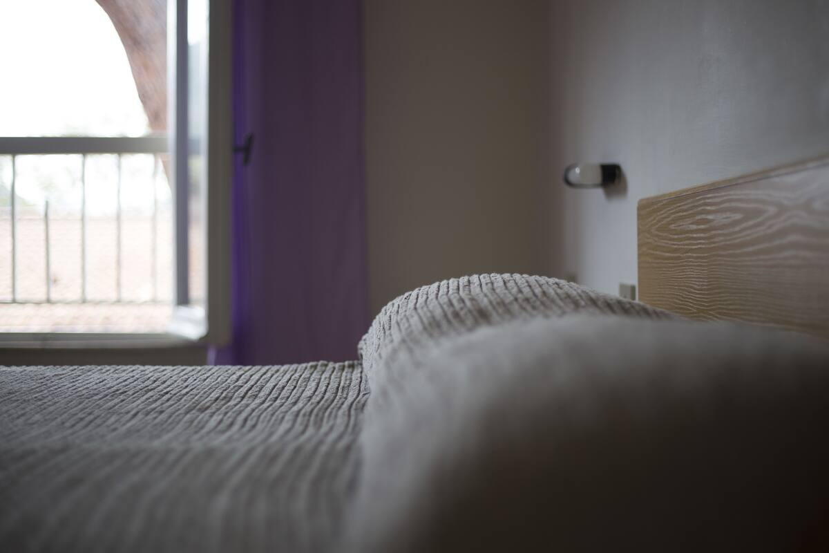 Primer plano de una cama con cubrecama blanco texturizado, cabecera de madera y una puerta con picaporte plateado.