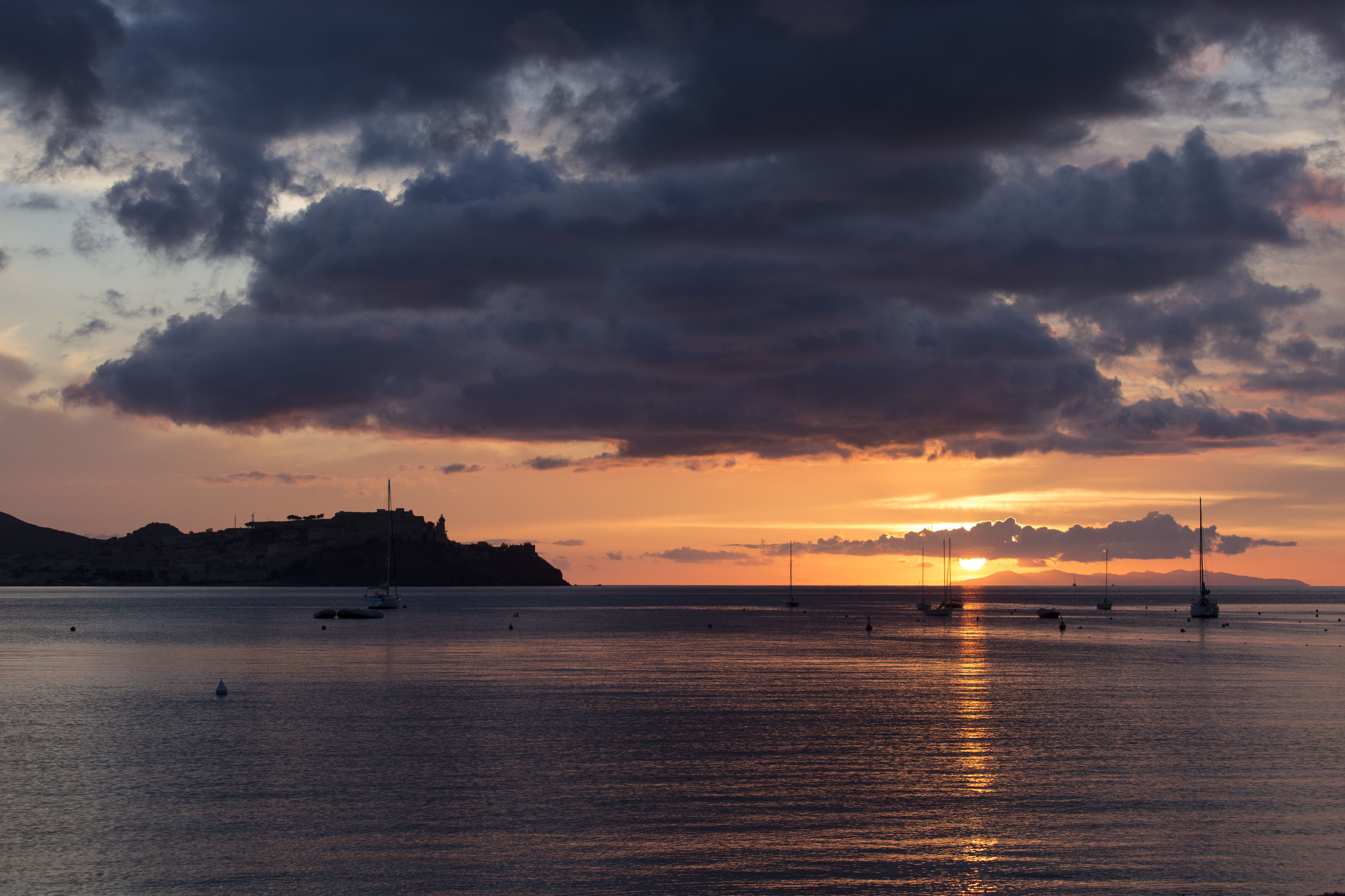 A sunset over a calm sea with boats and a distant hill.