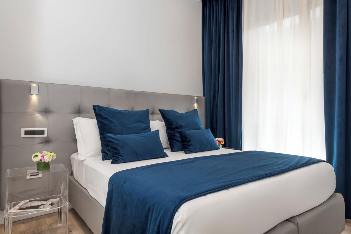 Un dormitorio moderno con cabecera gris, ropa de cama blanca y detalles azules.