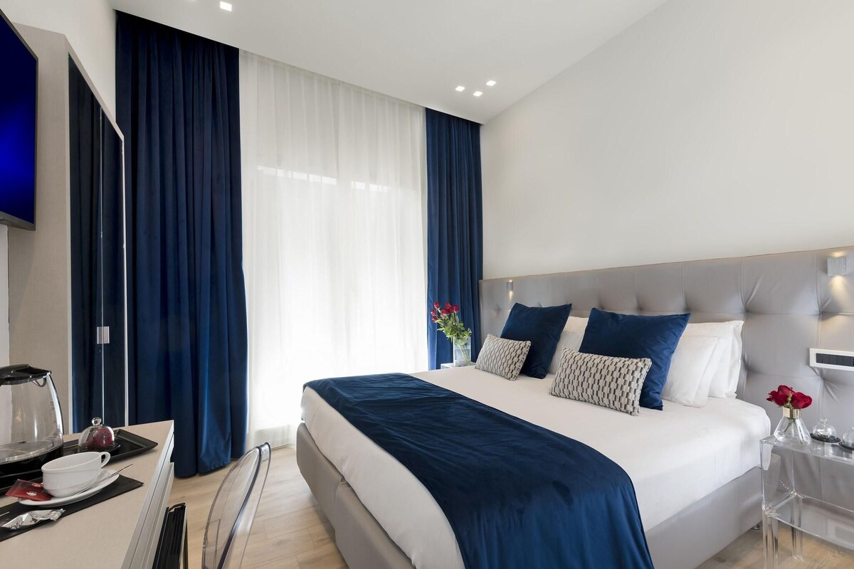 Una habitación de hotel moderna con una cama grande, una mesita de noche con un florero y un escritorio con cafetera.