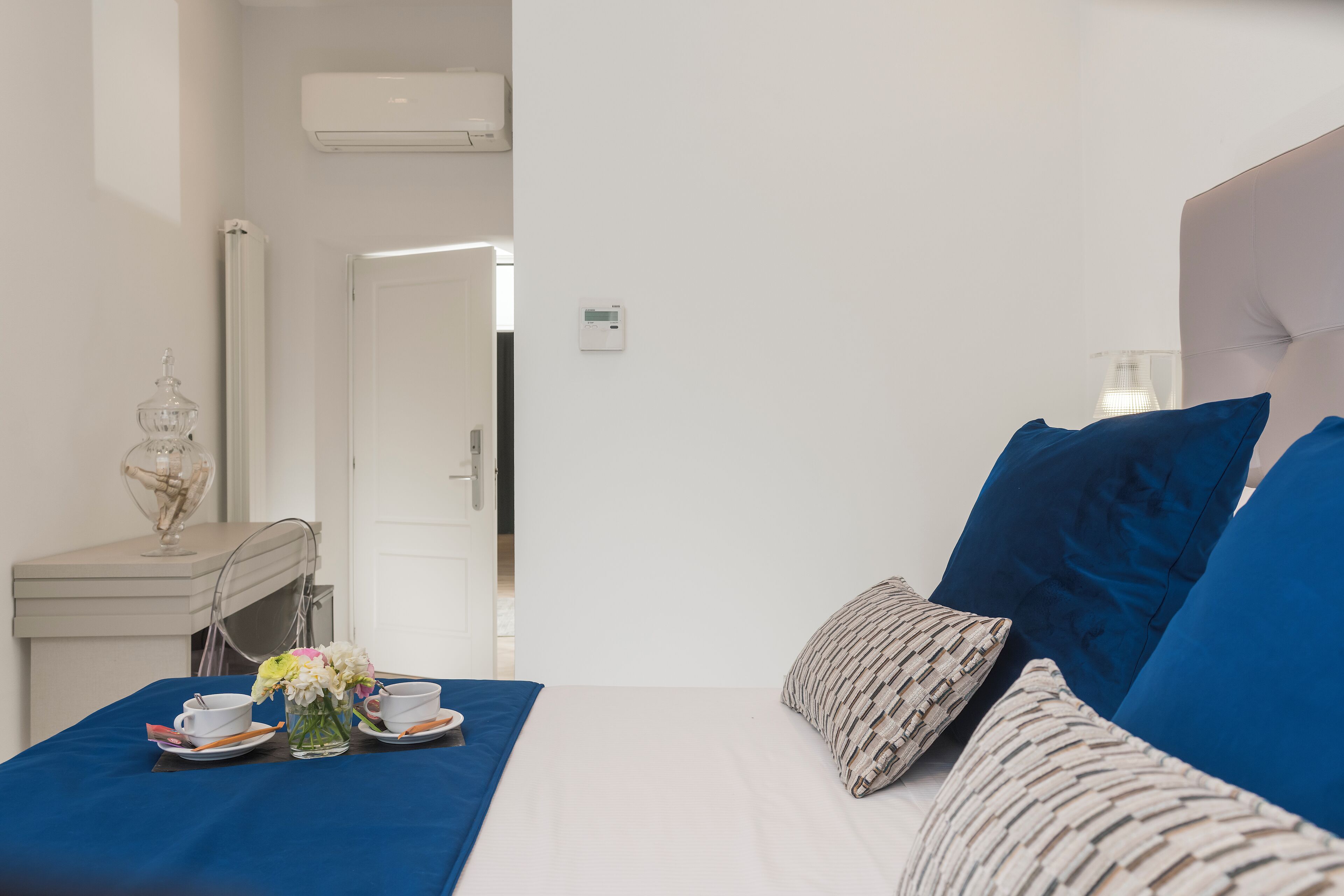 A hotel room with a bed, a bedside table with a vase of flowers, a cup and saucer, and a small table with a plate and a cup.