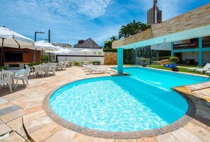 A swimming pool with lounge chairs and umbrellas, surrounded by a patio area with a stone floor. - Hotel Alimar (Natal)