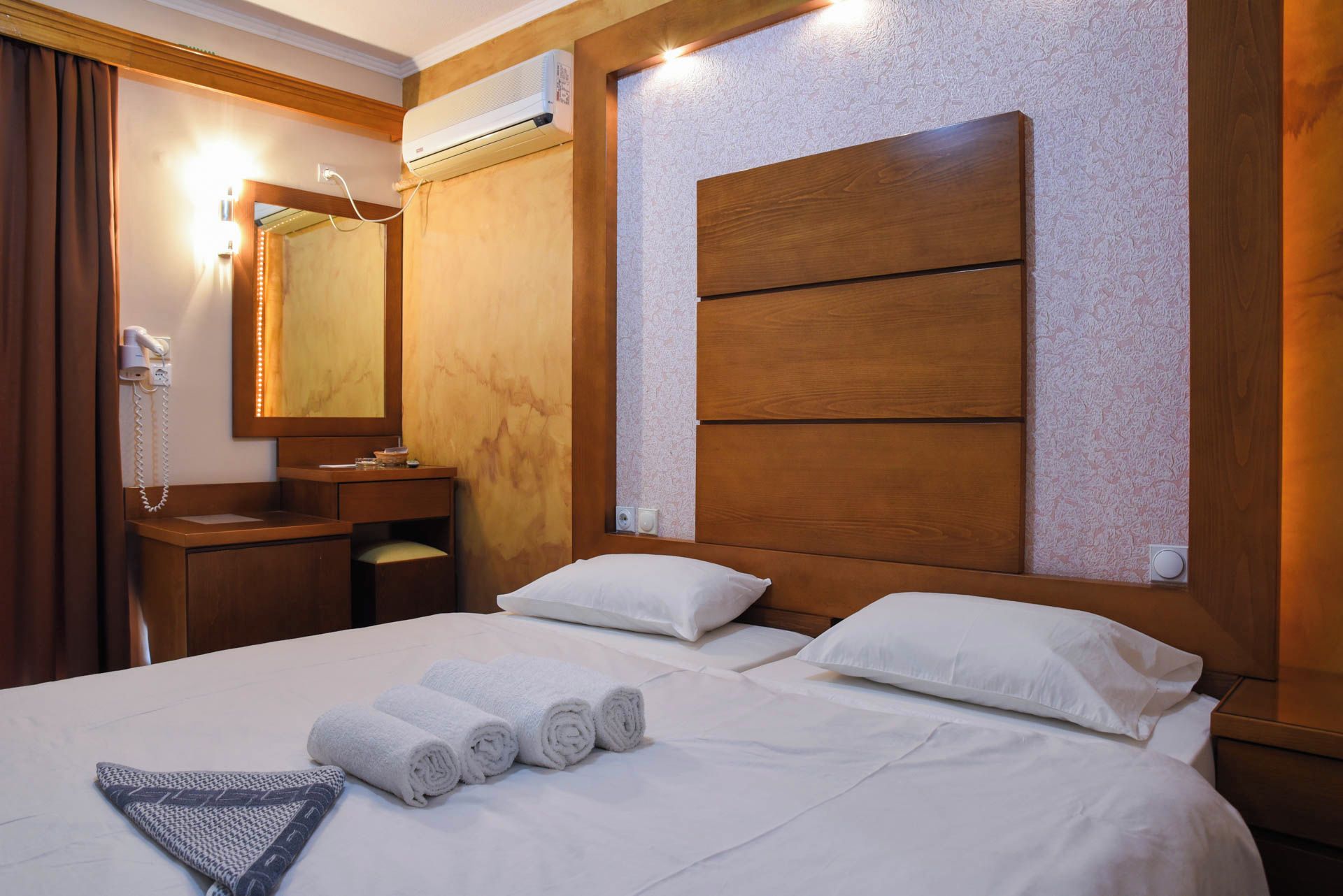 A hotel room with a bed, a wooden headboard, a bedside table with a lamp, a mirror, and an air conditioning unit.