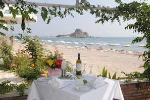 Een buitenterras met een tafel voor twee, met uitzicht op een strand met parasols en een rotsachtig eiland in de verte.
