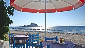 Een tafel aan het strand met een tafelkleed, blauwe stoelen en een rood-witte parasol.