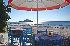 A beachside table with a tablecloth, blue chairs, and a red and white umbrella. - Sacallis Inn Beach Hotel (Kos)