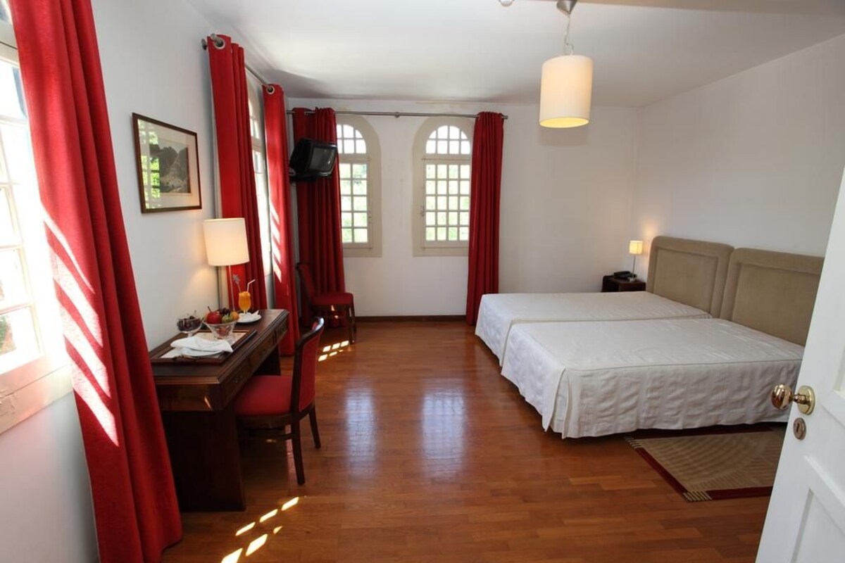 Une chambre à coucher avec un lit, un bureau et une chaise. Il y a deux fenêtres avec des rideaux rouges, un téléviseur fixé au mur et une lampe sur le bureau.