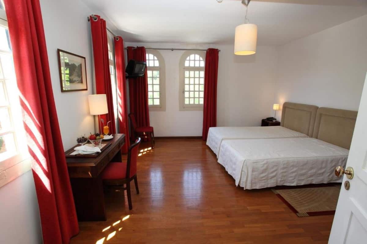 Une chambre avec un lit, un bureau et une chaise, une télévision et deux fenêtres avec des rideaux rouges.