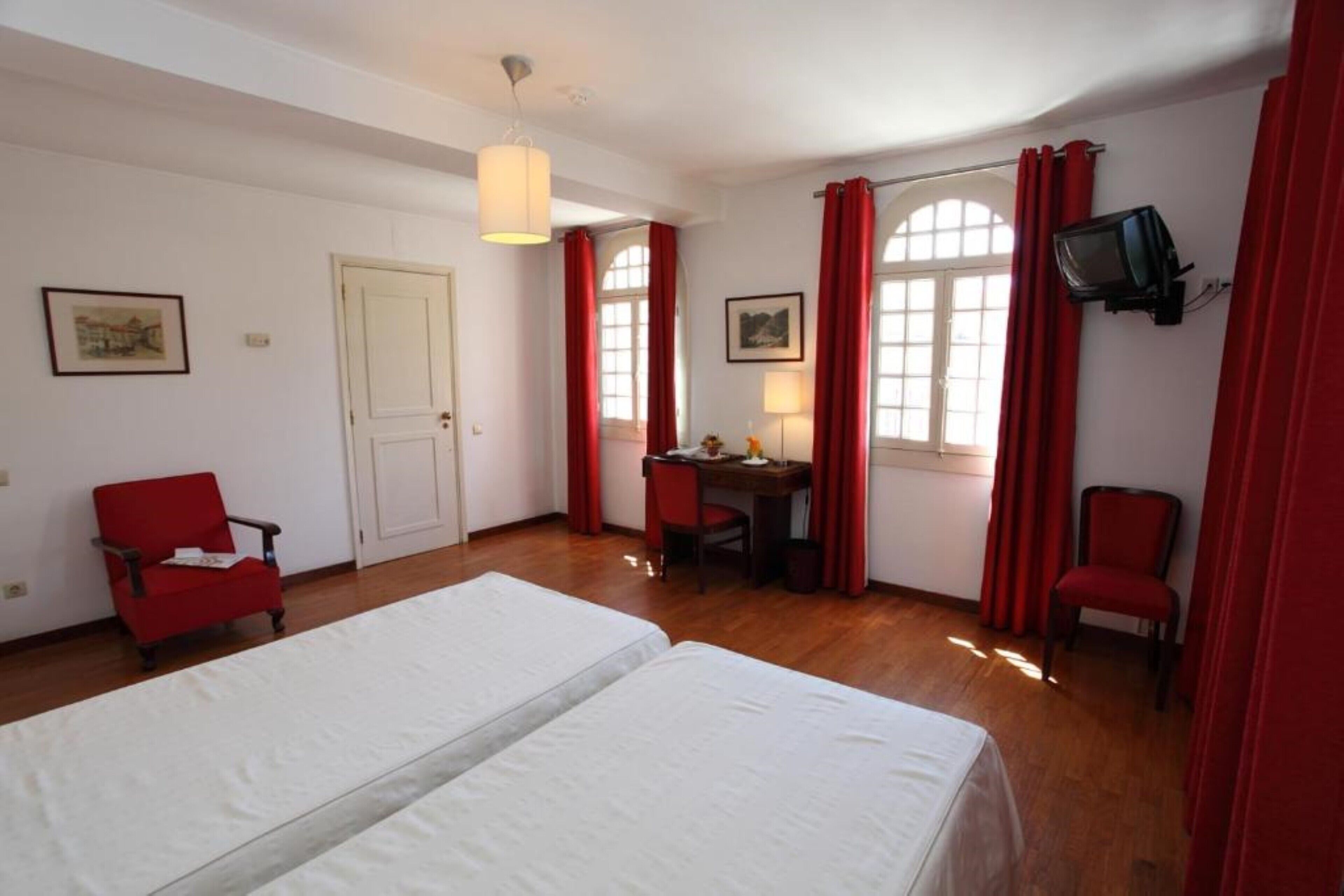 A room with two beds, a desk, a chair, a television, and red curtains.