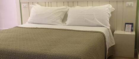 Una cama bien tendida con sábanas blancas y una manta gris, un cabecero blanco y dos lámparas de noche.