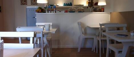 A dining area with white tables and chairs, a kitchen counter with food items, and a refrigerator.