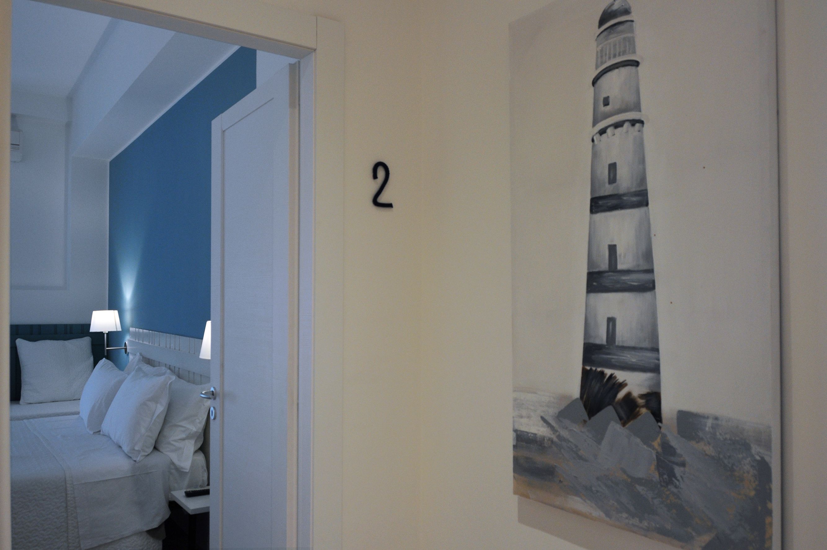 A hotel room with a bed, two bedside lamps, and a painting of a lighthouse on the wall.