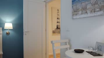 A room with a blue wall, a white door, a white chair, a small table with a bowl and glasses, and a painting of beach chairs on the wall.
