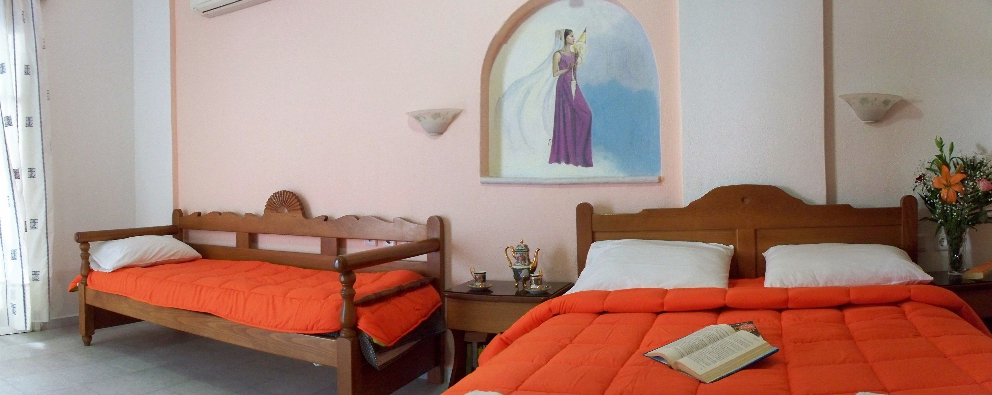 A hotel room with two wooden beds, each with an orange bedspread and white pillows. There is a small table with a decorative tea set and a wall-mounted picture of a woman in a purple dress.