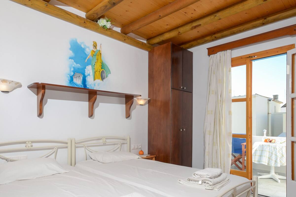A double bed with white linens, a wooden ceiling, a painting on the wall, and a view of the outside through a glass door.