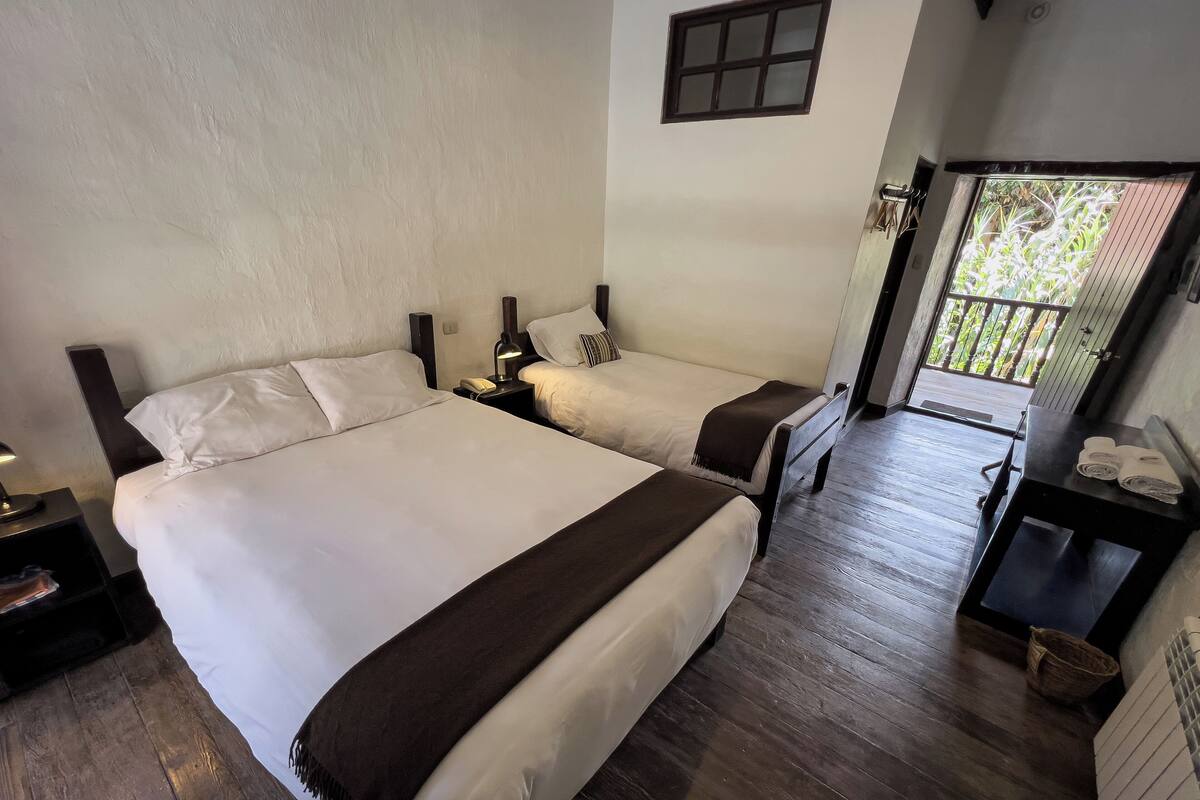 Een hotelkamer met twee bedden, houten vloeren, een klein tafeltje met handdoeken en een balkon met uitzicht op groen.